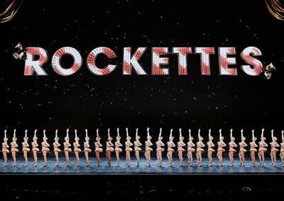 The word "rockettes" in red and white stripes on a black background above a line of dancers on a stage